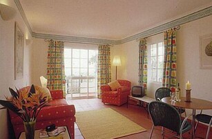 A living room with a red sofa, a small table with a lamp, and a chair. There are curtains on the windows and a vase with flowers on the sofa.