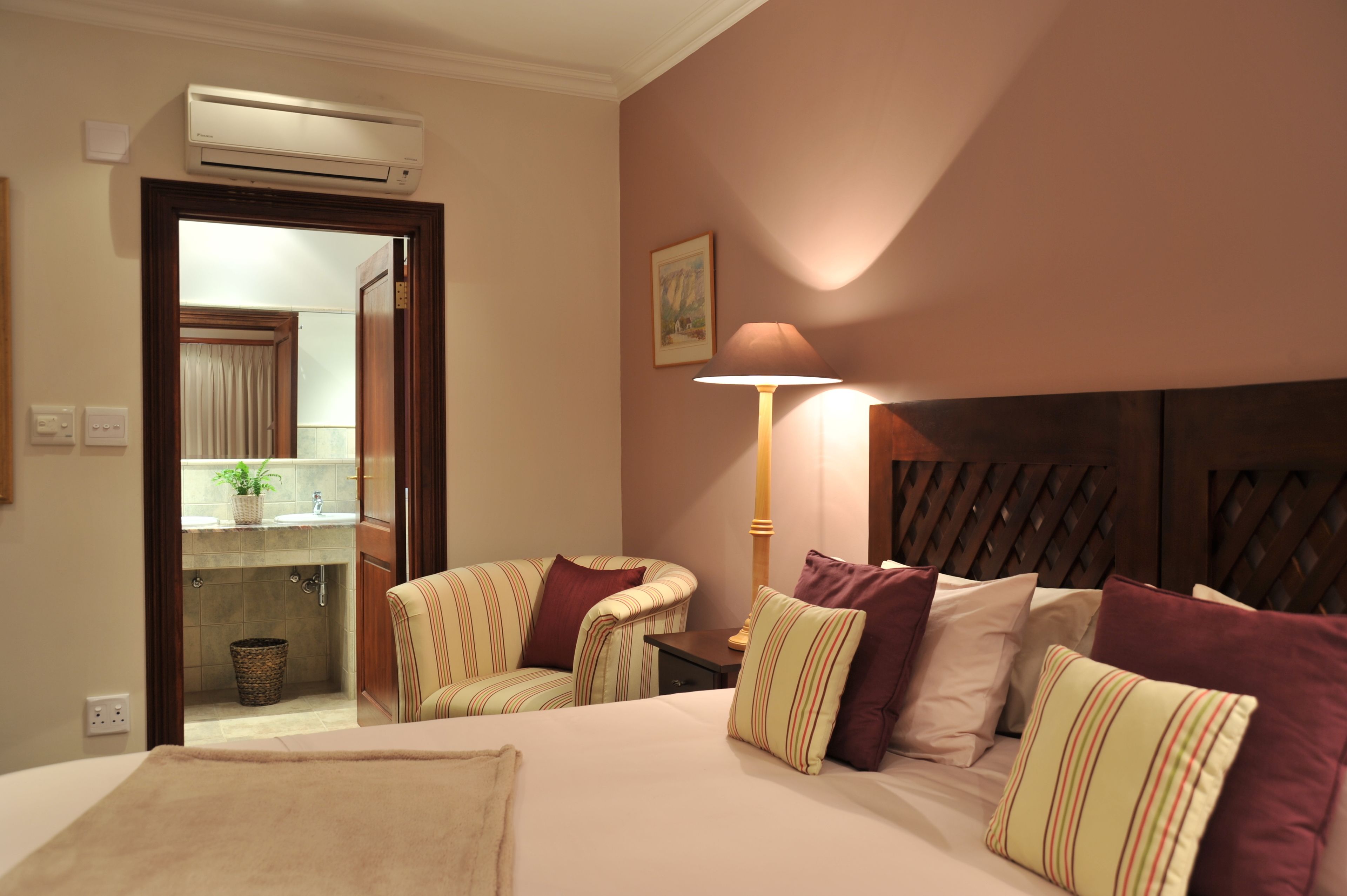 Habitación de hotel con cama, dos sillones, una mesita de noche, una lámpara, aire acondicionado de pared y un cuadro, con vistas al baño a través de una puerta abierta.