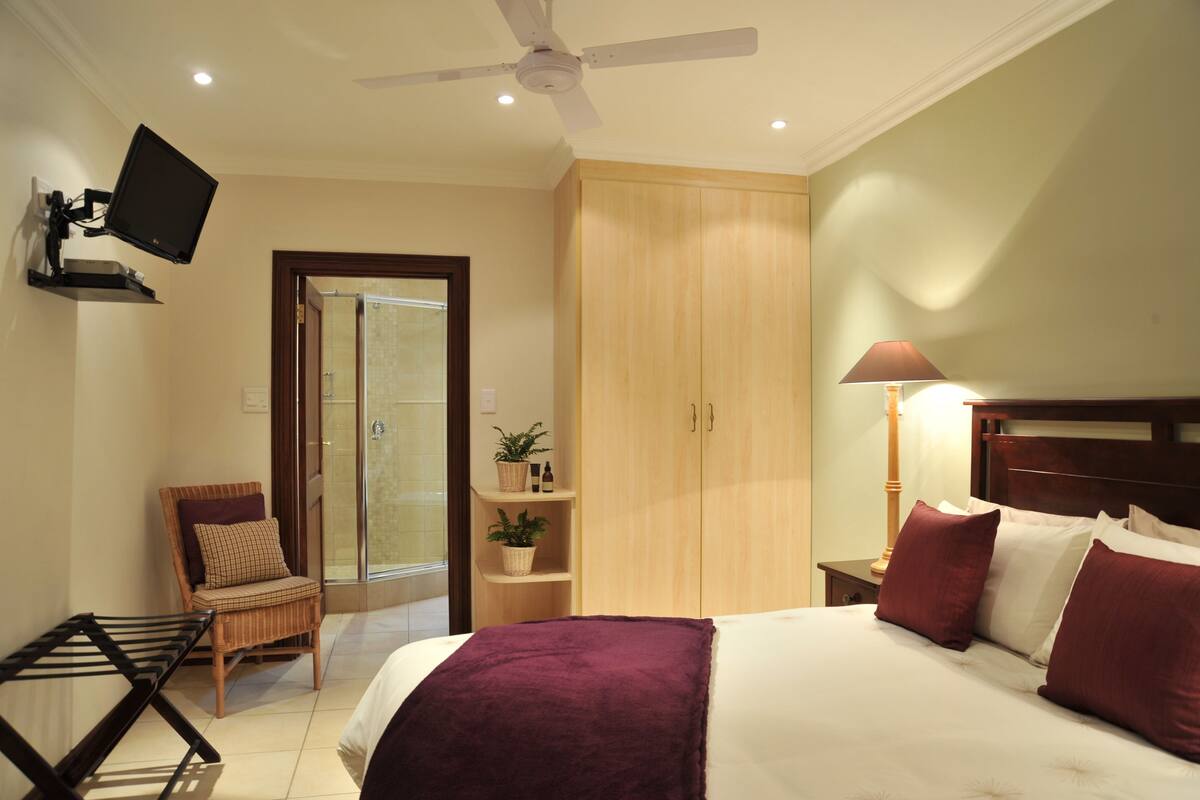 Una habitación de hotel con una cama, televisión montada en la pared, ventilador de techo, armario de madera, una mesita con lámpara y una silla.