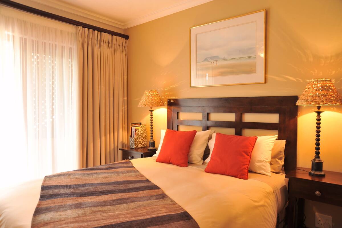 Habitación de hotel con una cama grande, dos mesitas de noche con lámparas, un cuadro enmarcado en la pared y una ventana con cortinas.