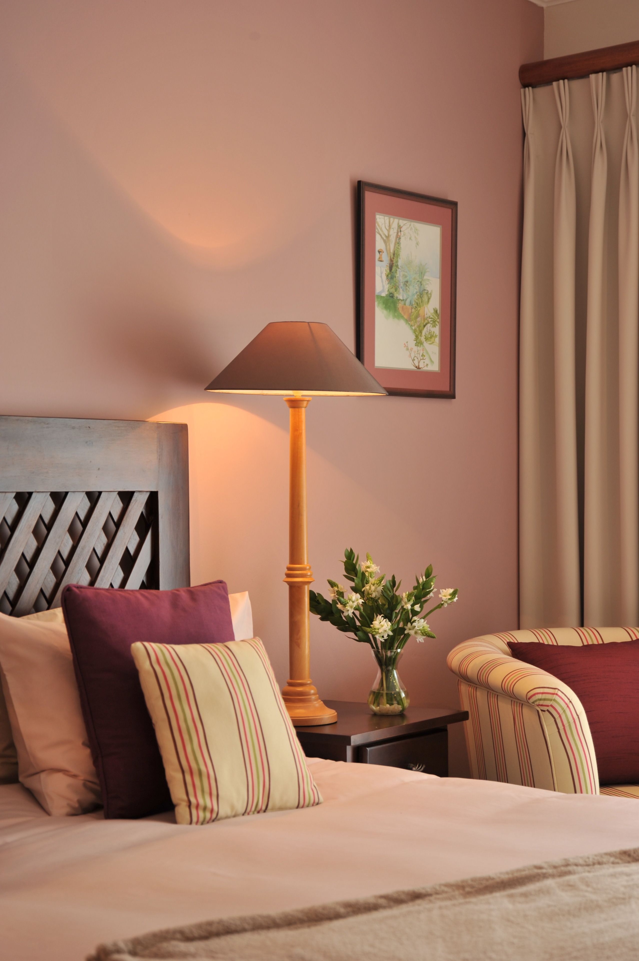 A neatly arranged hotel room with a bed, a bedside table with a lamp, a framed picture on the wall, and a window with curtains.
