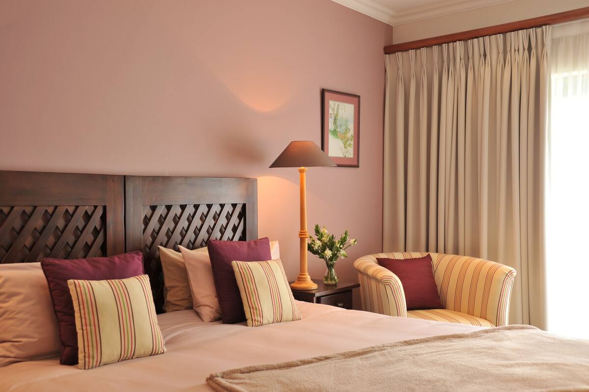 Habitación de hotel con cama, mesita de noche, lámpara, silla y ventana con cortinas.