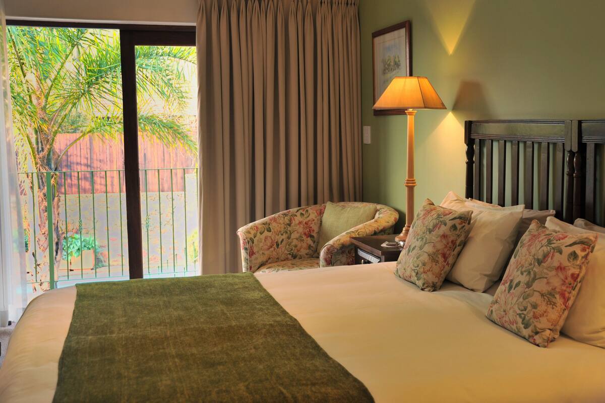 Habitación de hotel con cama, sillones estampados, lámpara y vistas a palmeras a través de la ventana.