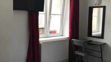 A hotel room with a bed, a desk, a chair, a window with curtains, a radiator, and a wall-mounted TV.