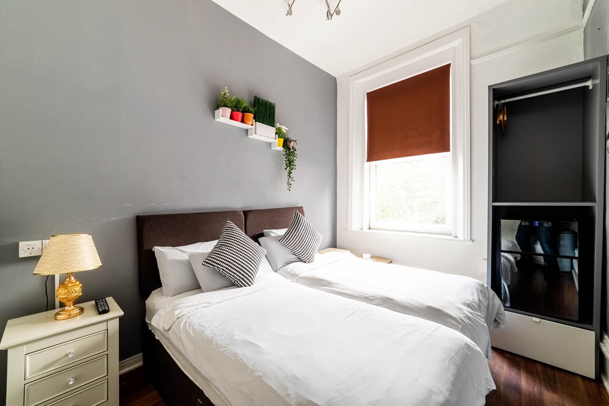A hotel room with two beds, a bedside table with a lamp, a shelf with plants, a window with a blind, and a wardrobe.