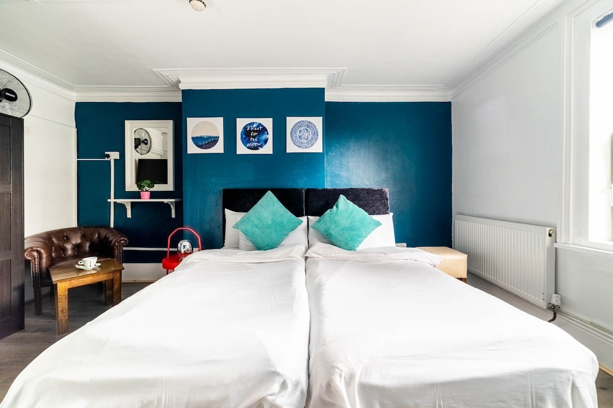 Quarto de hotel com uma cama grande, uma mesa pequena com uma xícara, uma cadeira e uma mala vermelha.