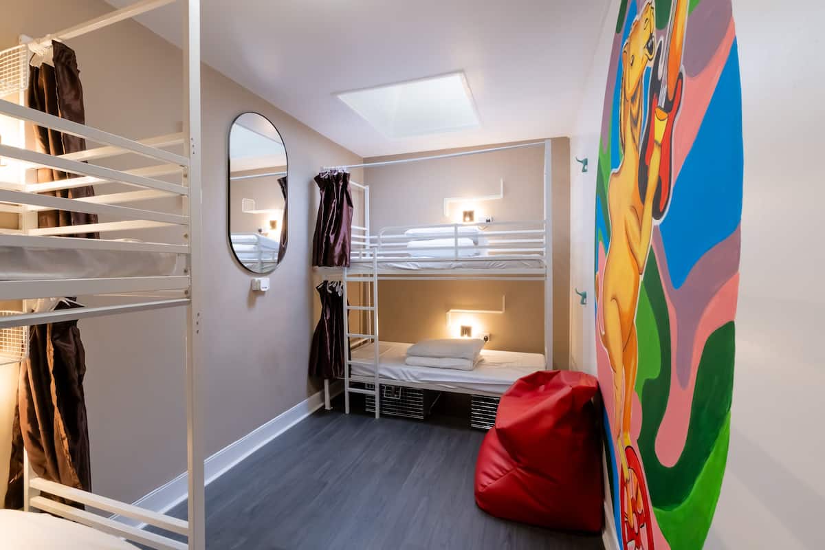 Une chambre avec un lit superposé, une lucarne, un pouf rouge et une fresque murale colorée.