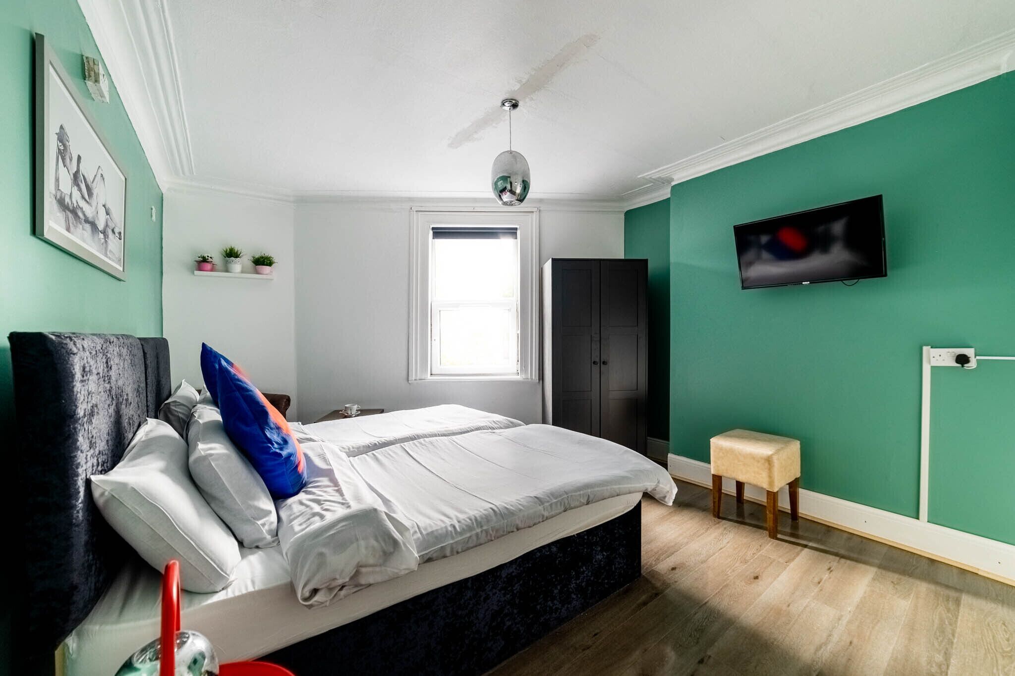 A bedroom with a bed, a TV mounted on the wall, a window, and a small stool.