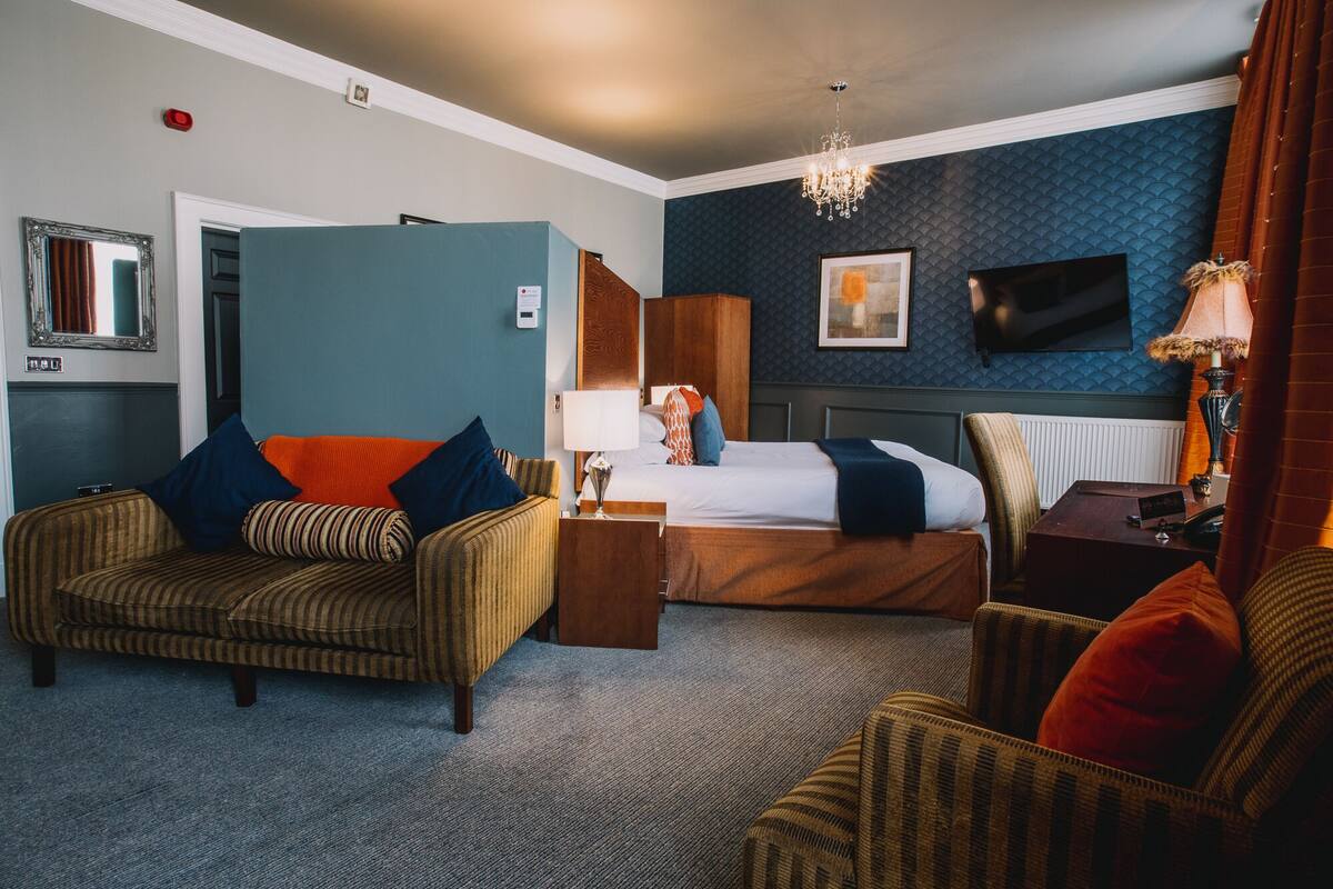 A hotel room with a bed, two armchairs, a nightstand, a TV, and a chandelier.
