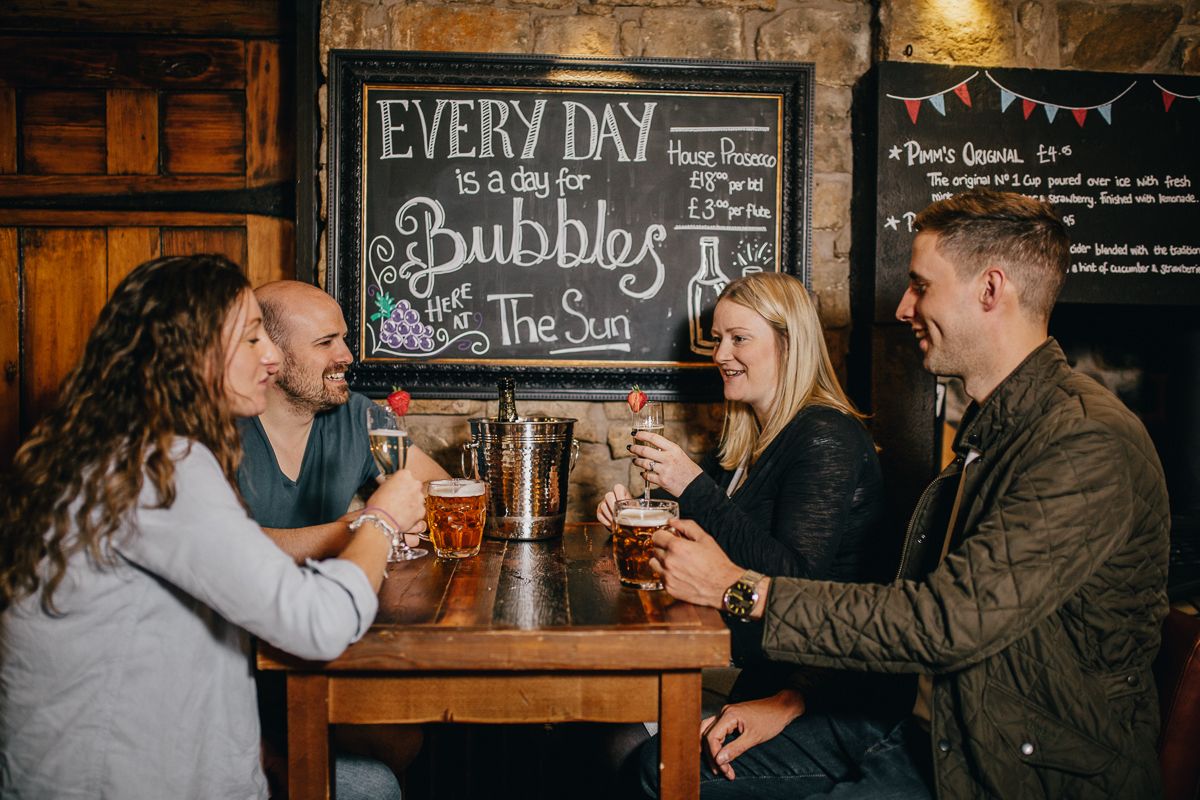 A group of people sitting around a wooden table with drinks, a chalkboard menu, and a stone wall in the background.