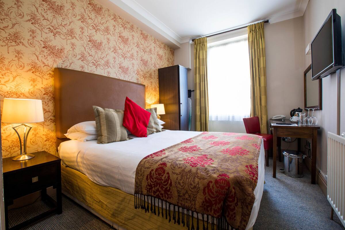 A hotel room with a large bed, a desk with a computer, a TV, and a window with curtains.