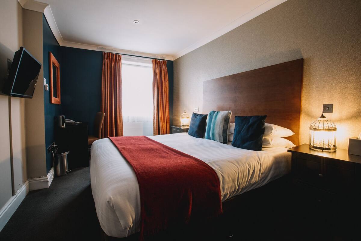 A hotel room with a large bed, a TV mounted on the wall, a bedside table with a lamp, and a window with curtains.