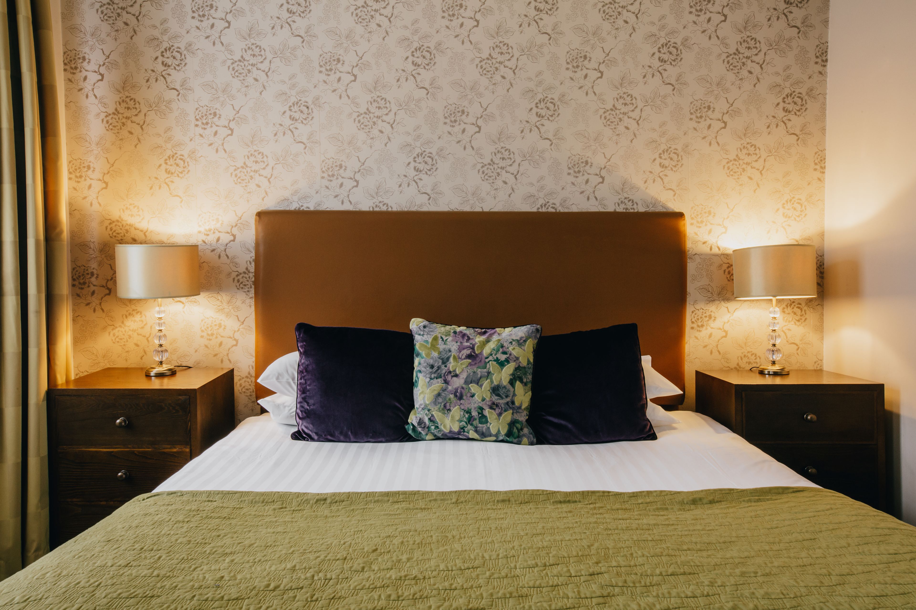 A neatly made bed with a green blanket, two dark pillows, and a floral throw pillow. There are two bedside lamps on wooden nightstands.