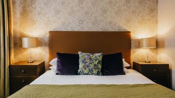 A neatly made bed with a green blanket, two dark pillows, and a floral throw pillow. There are two bedside lamps on wooden nightstands.