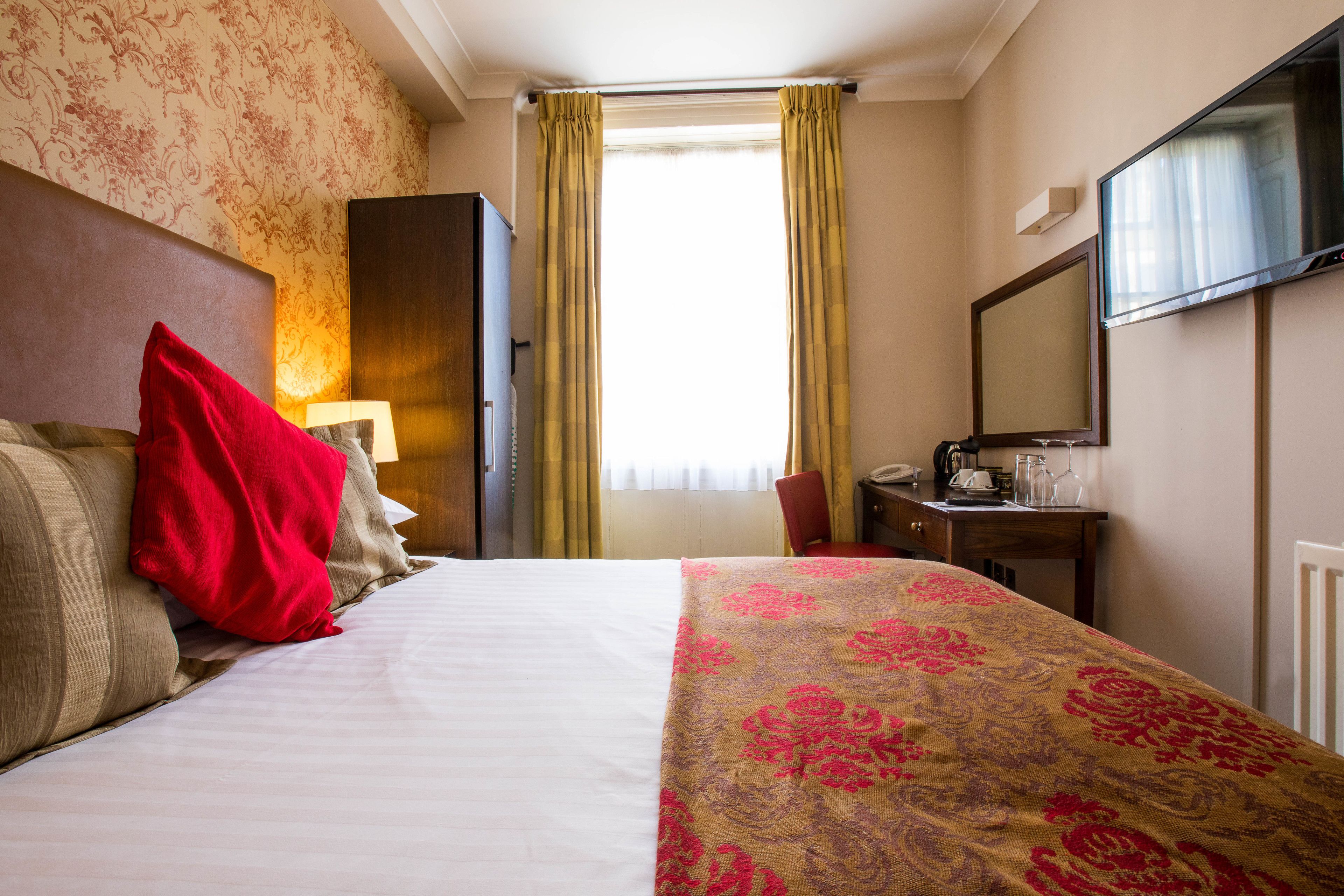 Quarto de hotel com uma cama, uma escrivaninha com telefone, um espelho e uma janela com cortinas.