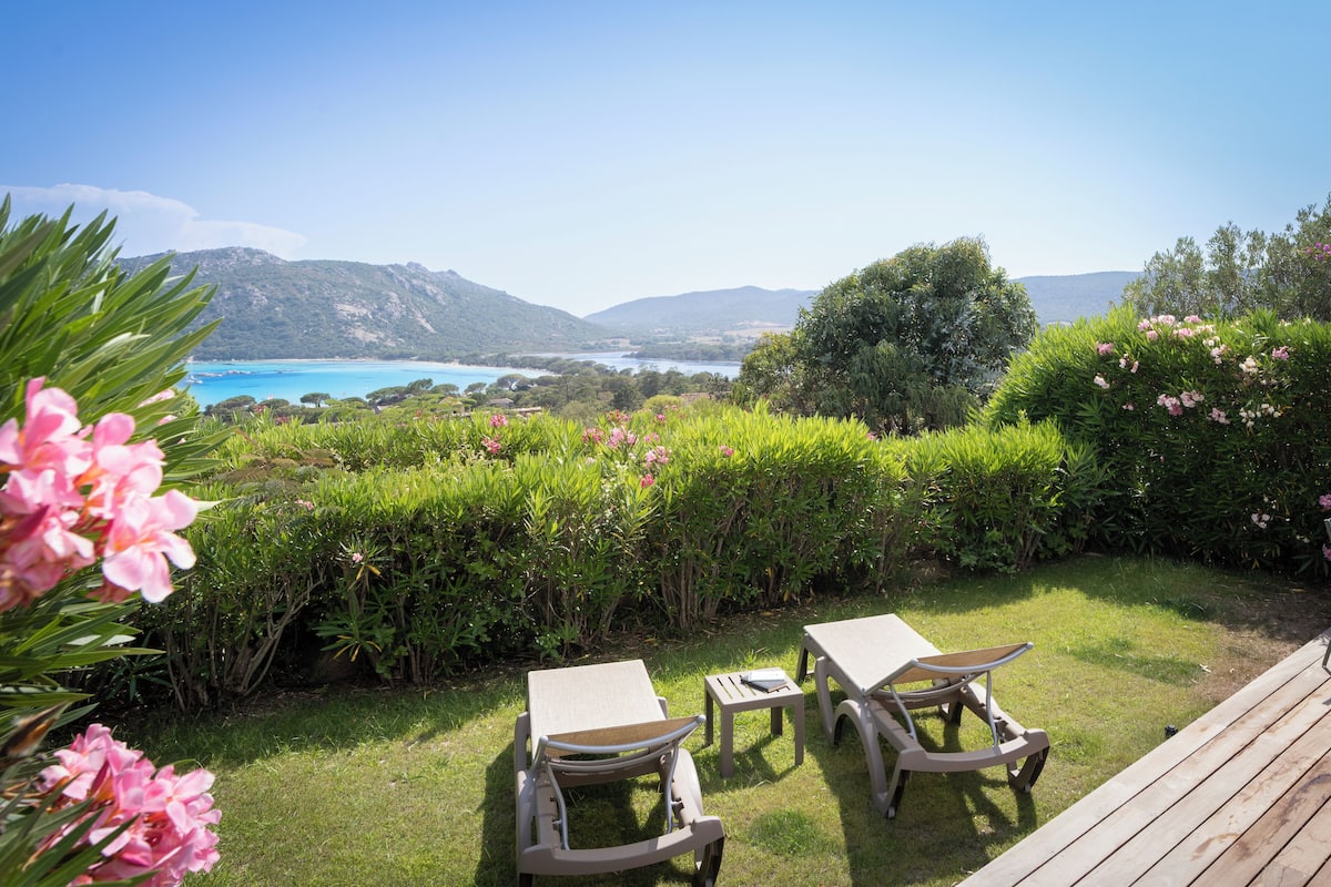 A garden with two tables and chairs, surrounded by lush greenery and a view of a lake and mountains.
