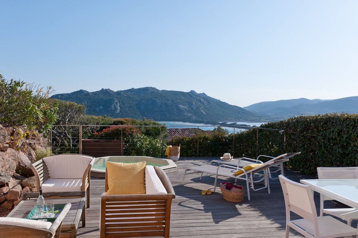 A wooden deck with outdoor furniture, a hot tub, and a view of the ocean and mountains.