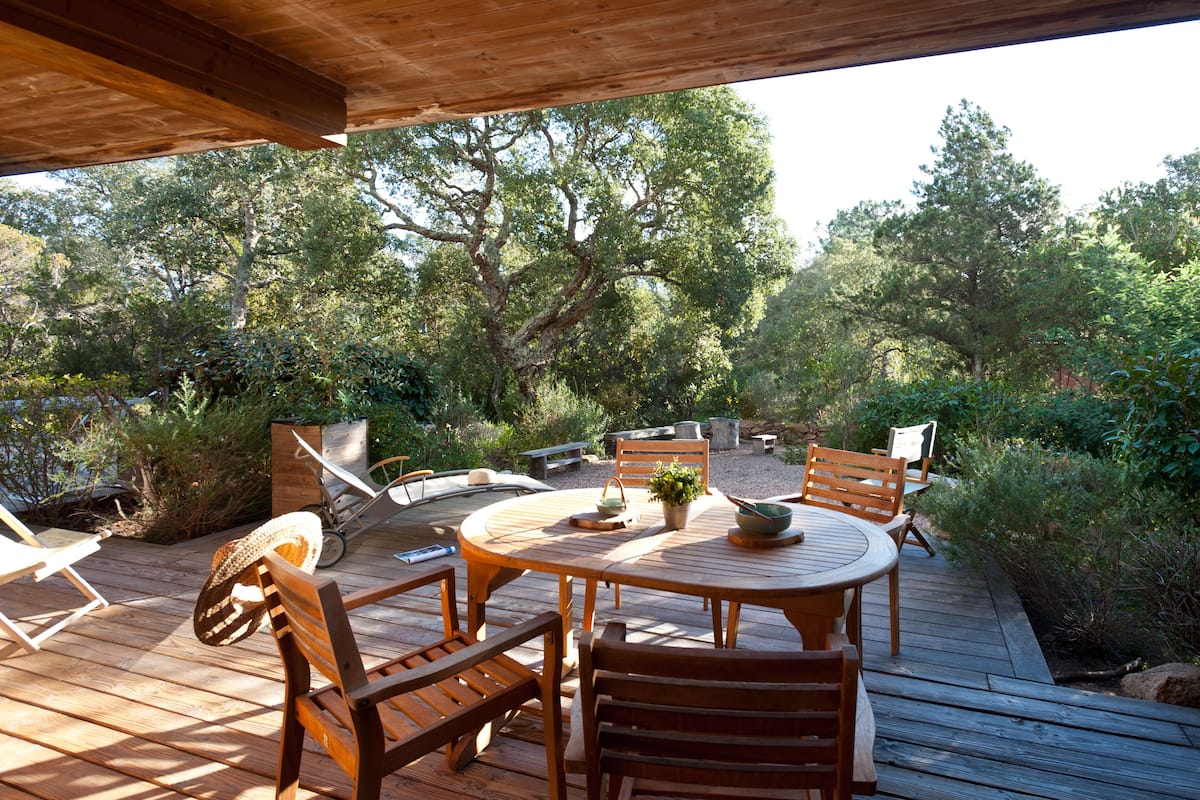 A wooden deck with a round table and chairs, surrounded by greenery and trees.