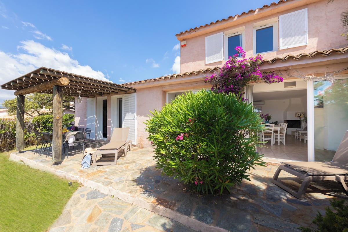 A house with a stone patio, a covered outdoor seating area, and a lush green bush with pink flowers.