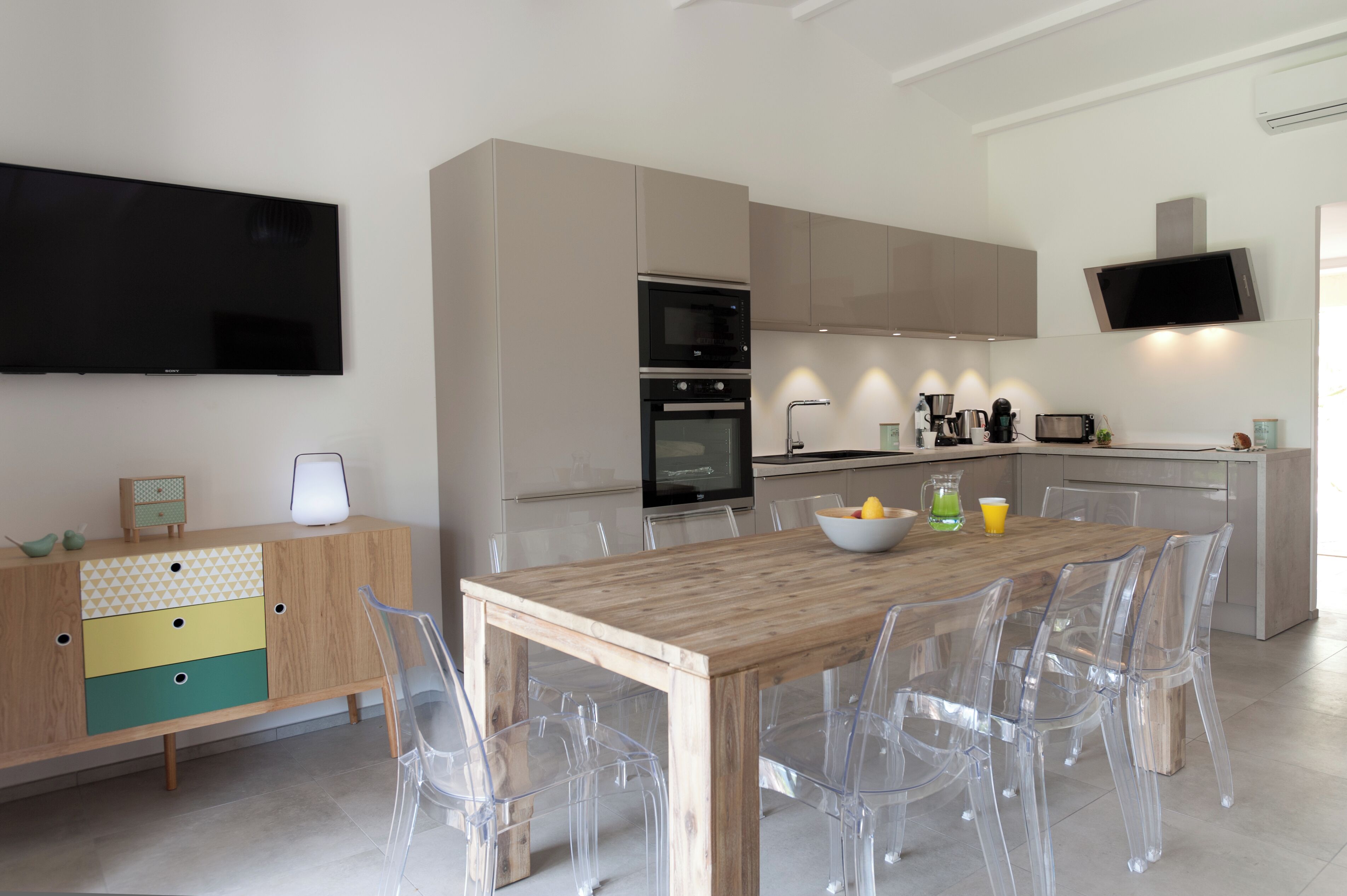 A modern kitchen with a wooden dining table, clear chairs, and a TV mounted on the wall.