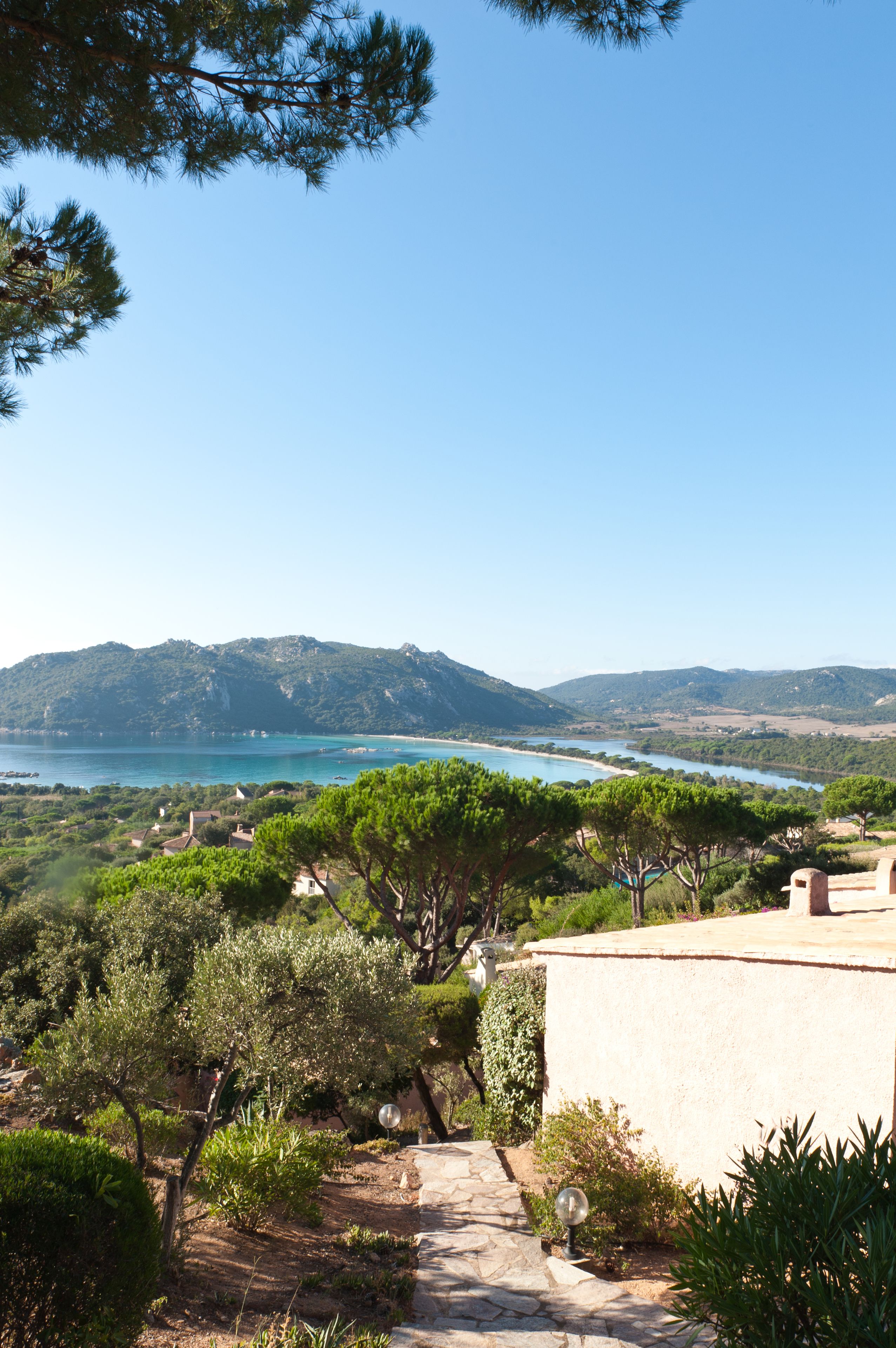 Un paysage côtier pittoresque, avec un sentier menant à un bâtiment, entouré de verdure et offrant une vue sur une baie, avec des montagnes à l’horizon.