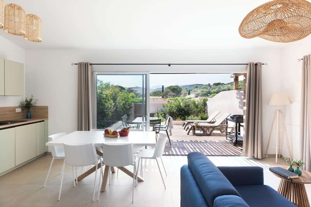A modern kitchen with white cabinets and a dining area with a white table and chairs. There is a blue sofa and a wicker hanging light fixture.
