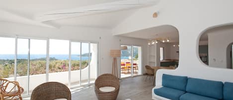 Amplia sala de estar con ventanas grandes, un sofá azul, sillas de mimbre y vistas al exterior.