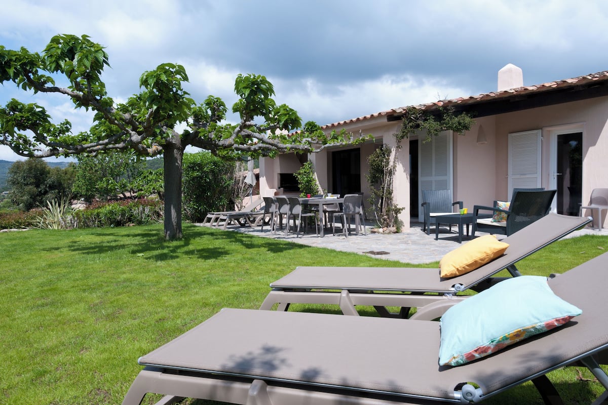 A patio with lounge chairs and a table, surrounded by a well-maintained lawn and a tree.