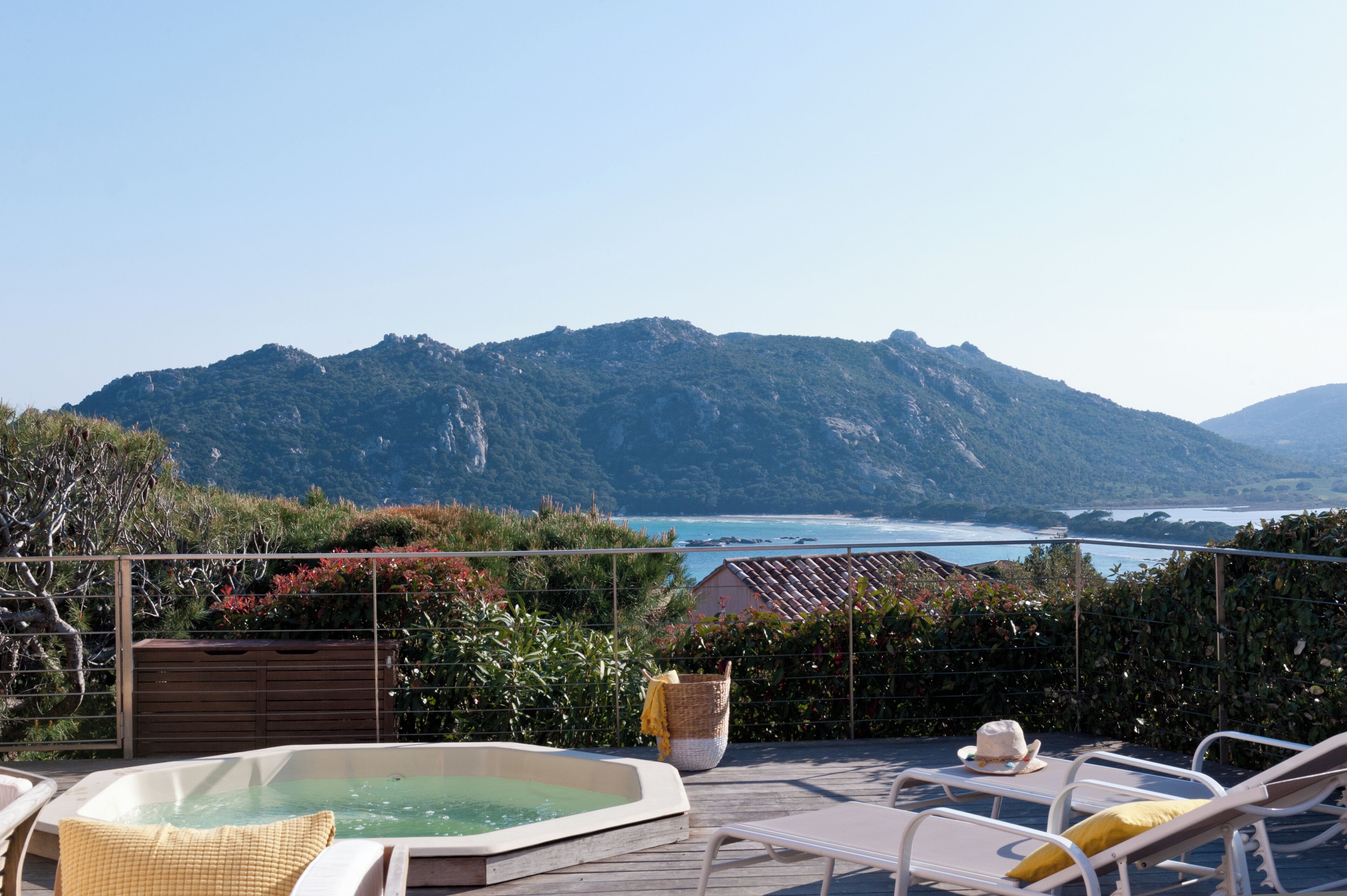 A hot tub on a wooden deck with lounge chairs and a view of a mountain and sea.