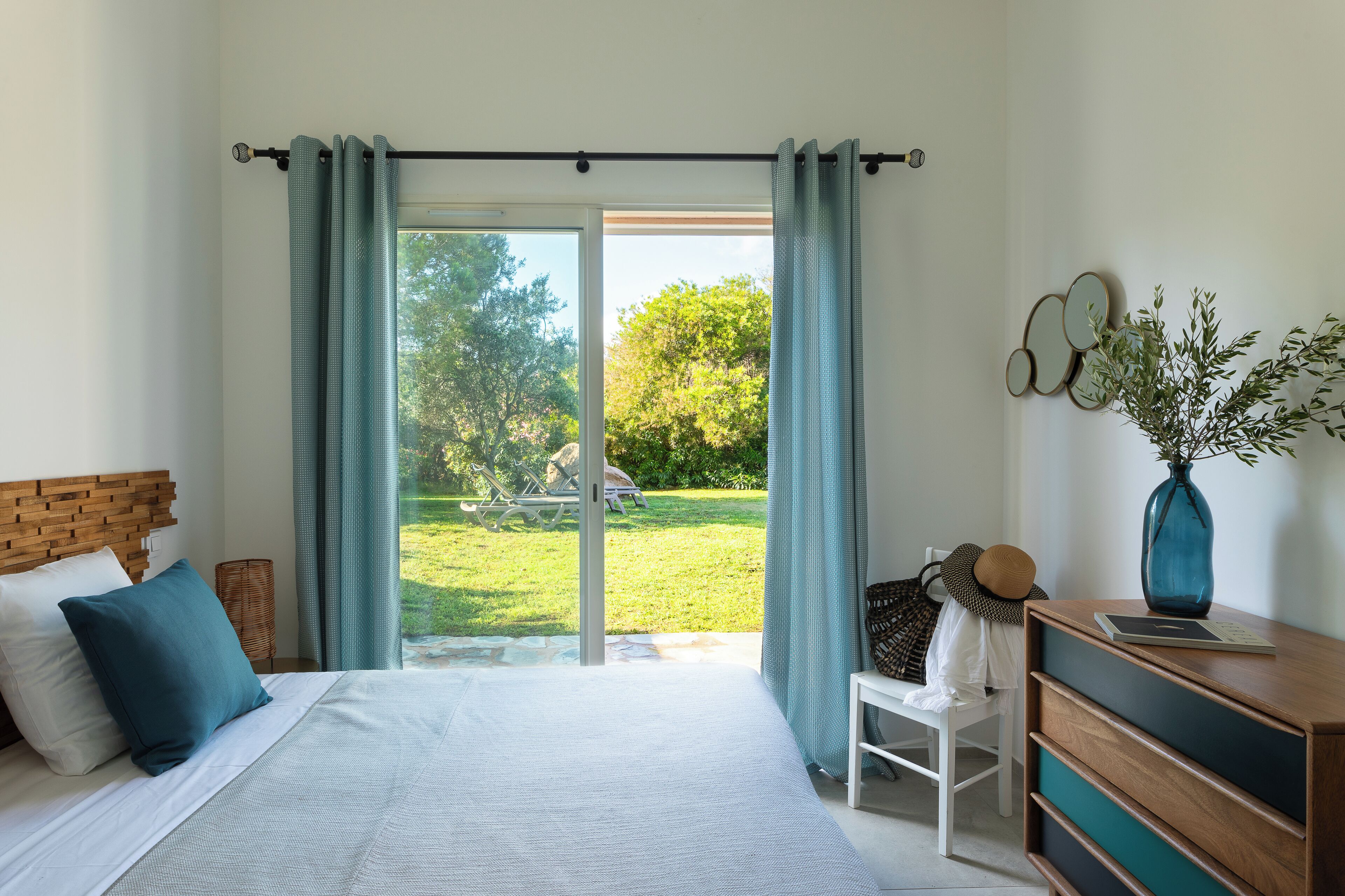 A bedroom with a bed, a chair, a dresser, and a view of a garden through a sliding door.