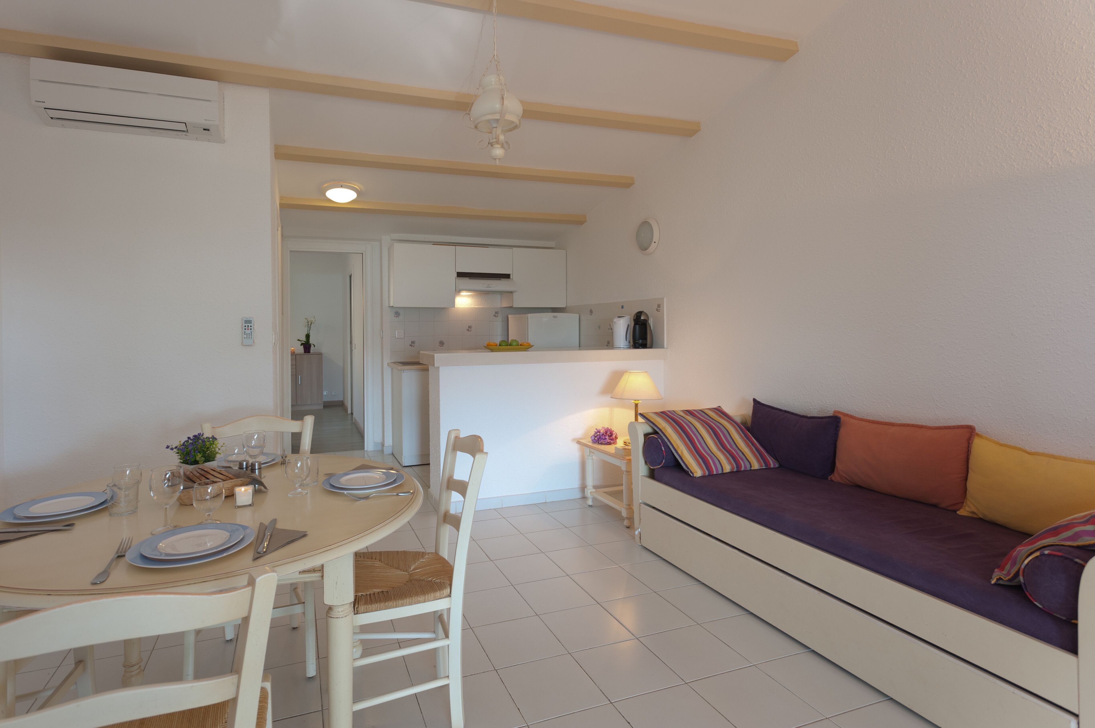 Un salon moderne avec une table à manger dressée pour quatre personnes, un canapé agrémenté de coussins colorés et une cuisine en arrière-plan.