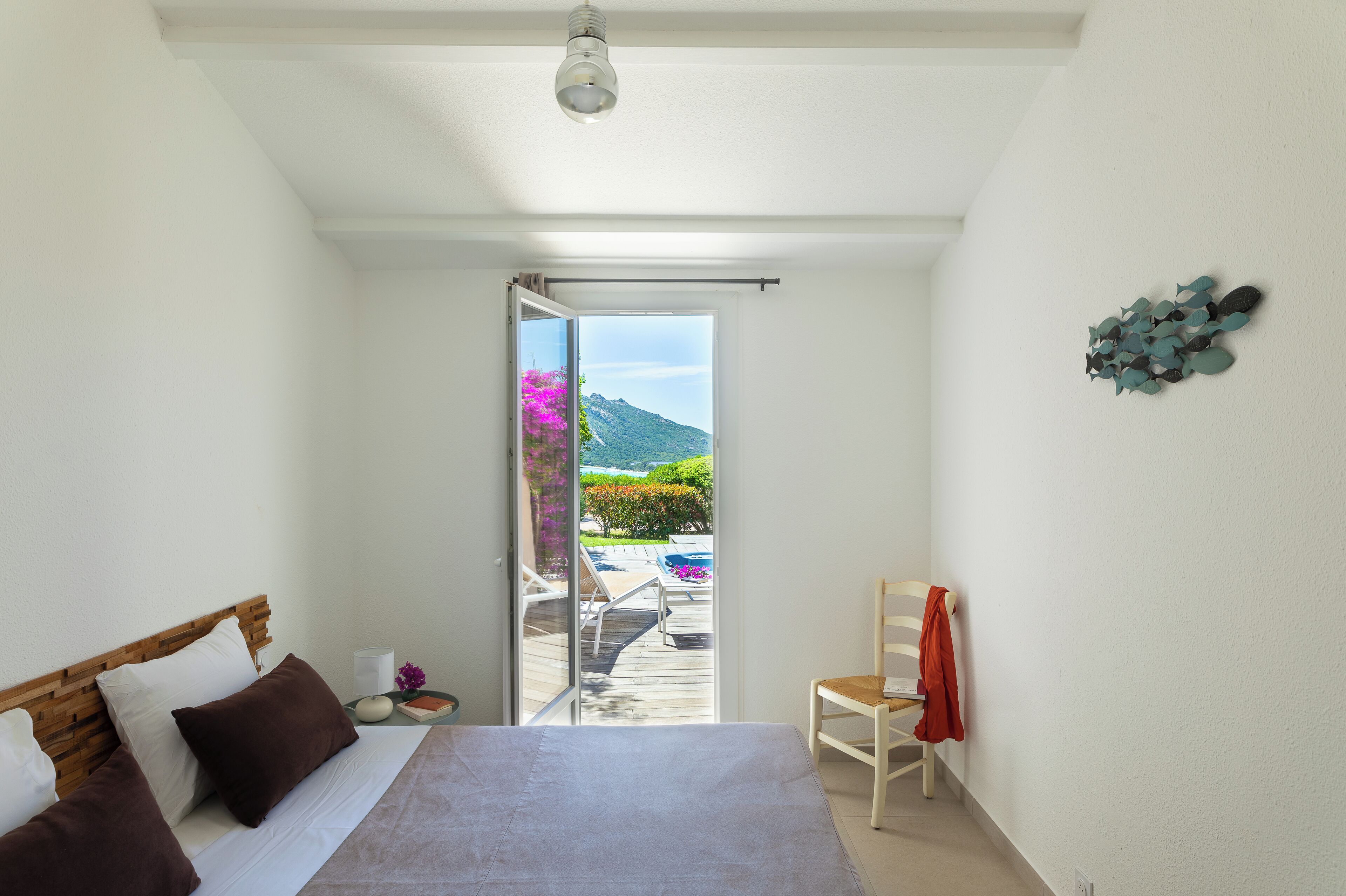 A bedroom with a bed, a chair, a nightstand, and a view of a balcony with outdoor furniture and a mountain in the distance.