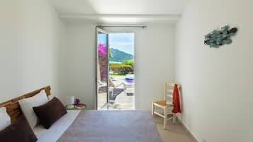 A bedroom with a bed, a chair, a nightstand, and a view of a balcony with outdoor furniture and a mountain in the distance.