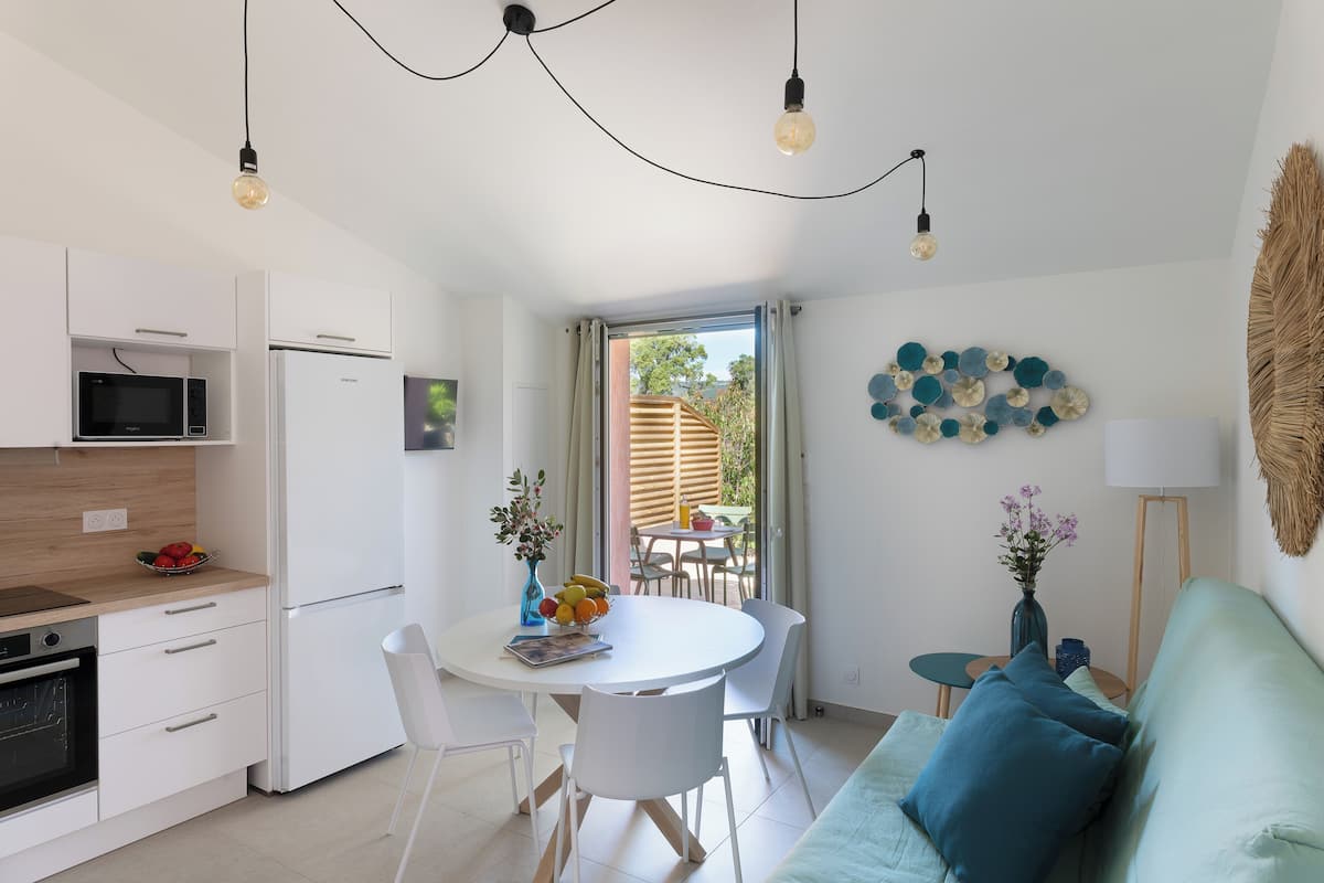 A modern kitchen with white appliances, a dining area with a white table and chairs, and a view of the outdoors through a glass door.