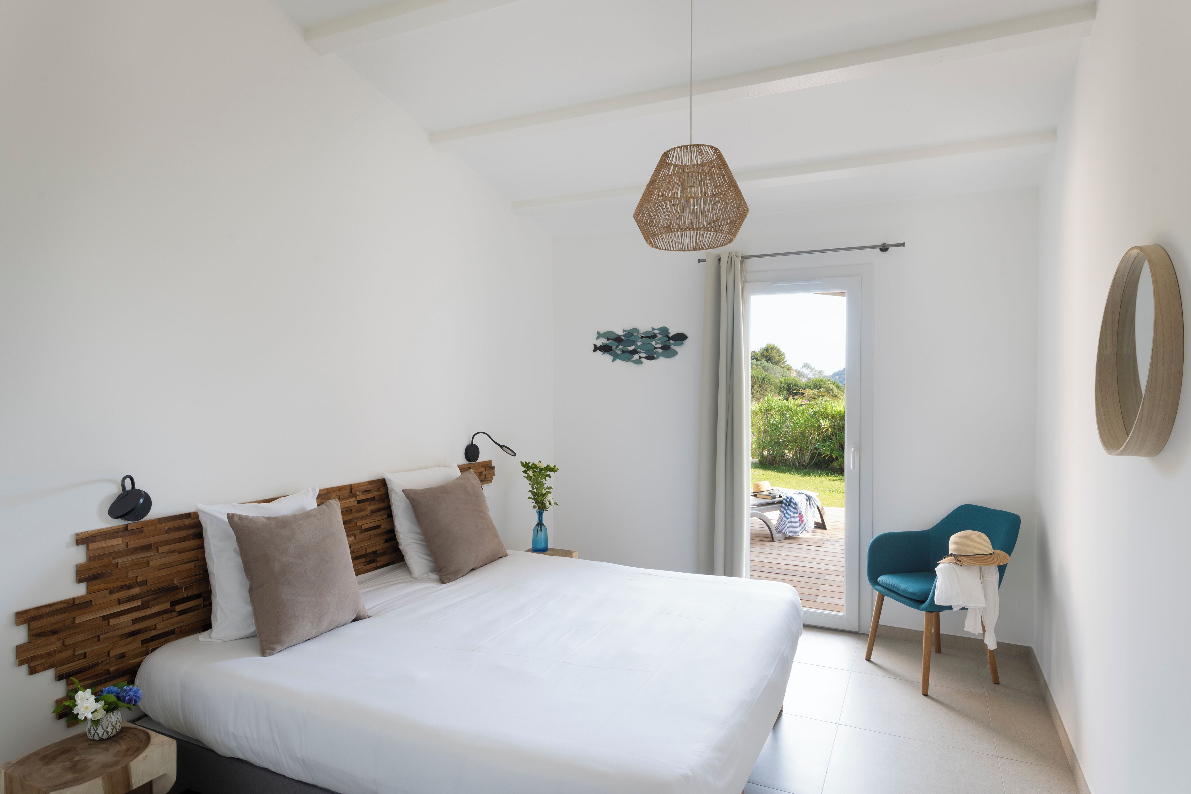 A bedroom with a large bed, a wooden headboard, a bedside table with a lamp, a vase with flowers, and a view of the outdoors through a sliding door.