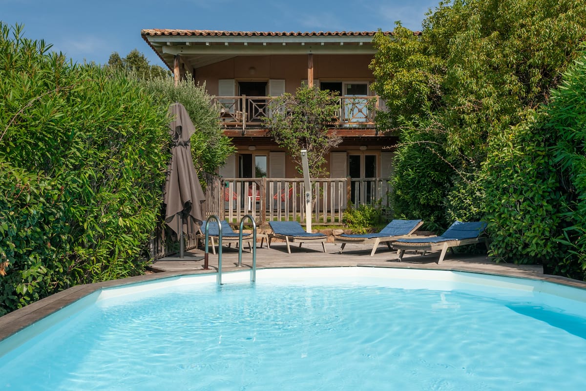 A swimming pool with lounge chairs and a covered area, surrounded by greenery and a building with balconies.
