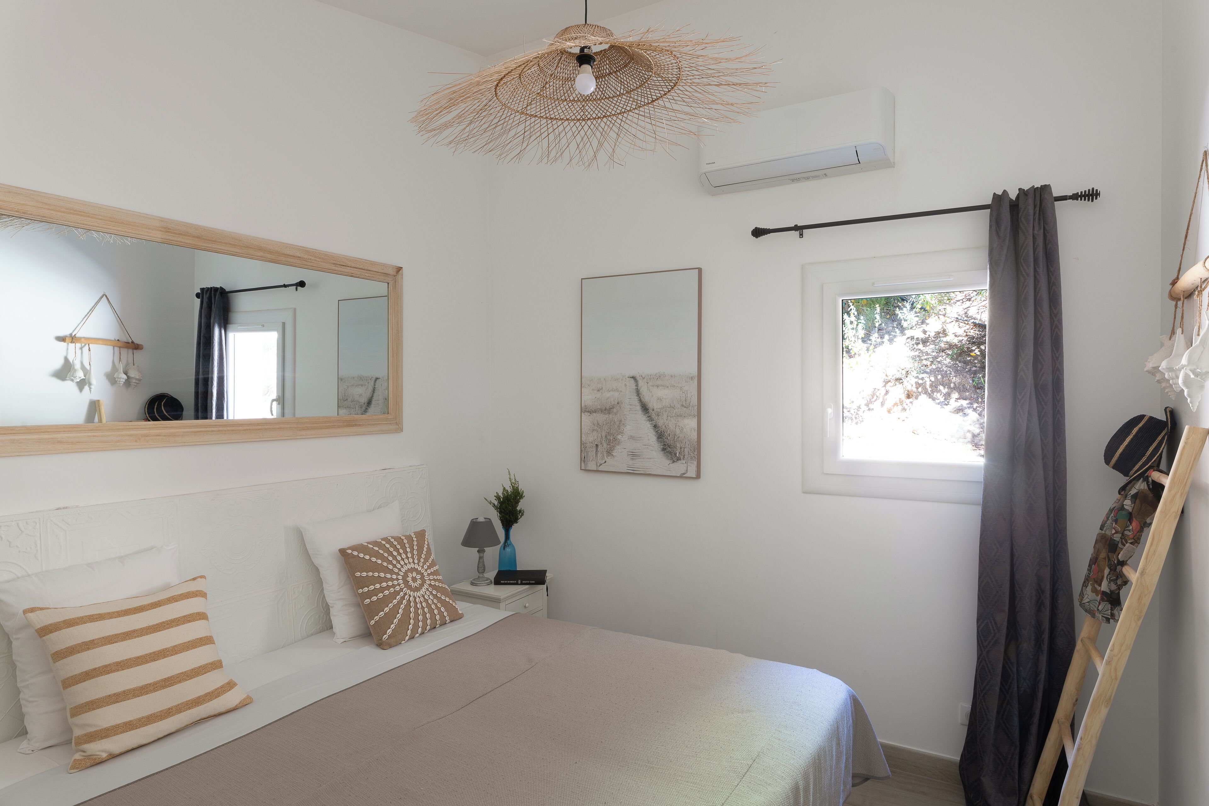 A bedroom with a bed, a bedside table, a mirror, a window with curtains, and a hanging light fixture.