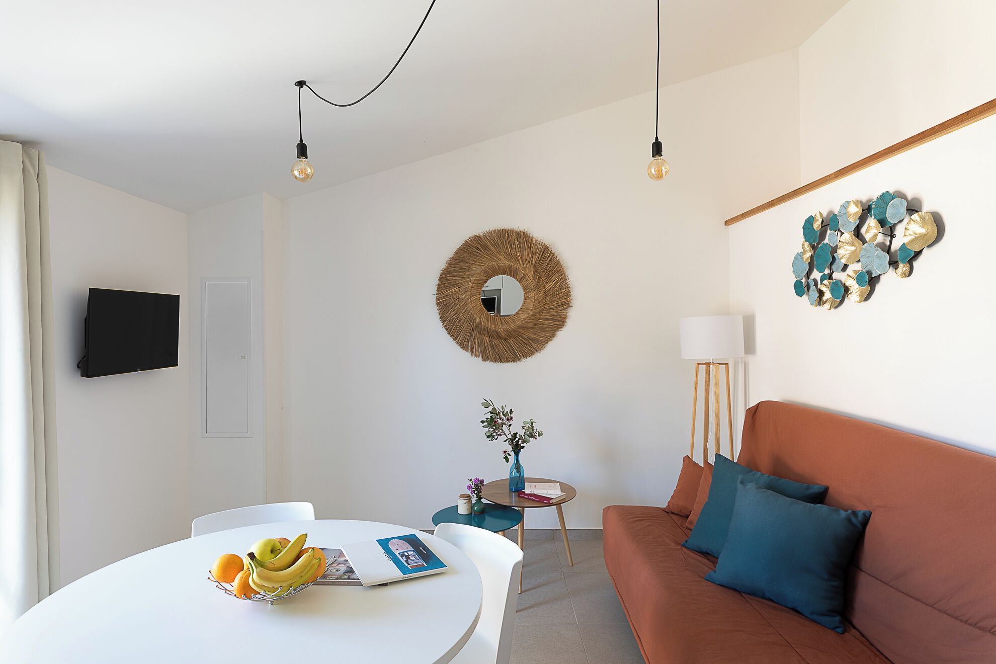 En moderne stue med et hvidt bord, en brun sofa, et rundt spejl og et tv ophængt på væggen.