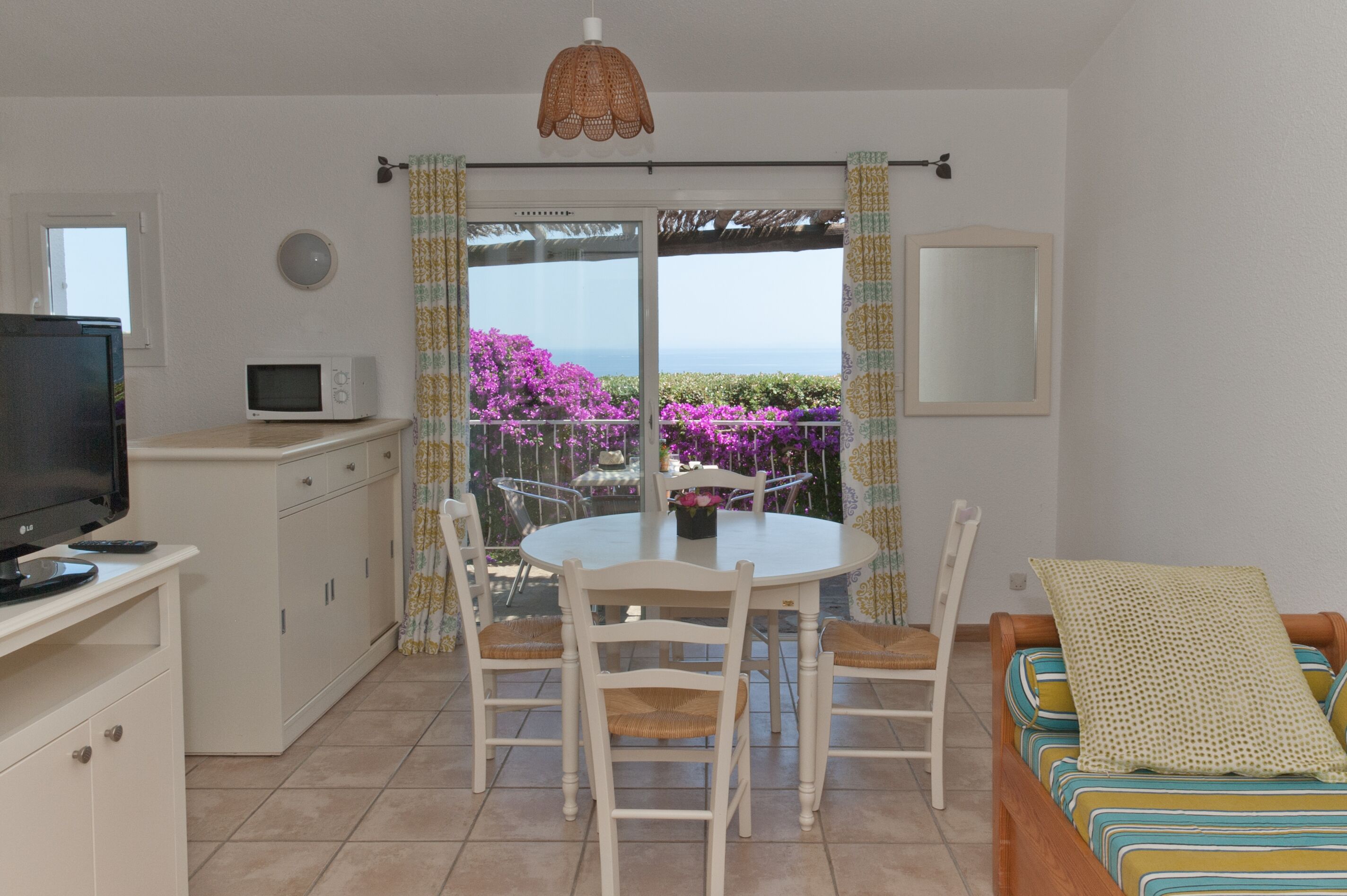 A compact living space with a kitchenette, a dining area with a table and chairs, a sofa, and a balcony with a view of purple flowers and the sea.