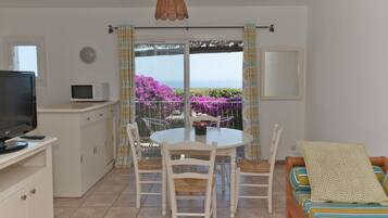 A compact living space with a kitchenette, a dining area with a table and chairs, a sofa, and a balcony with a view of purple flowers and the sea.