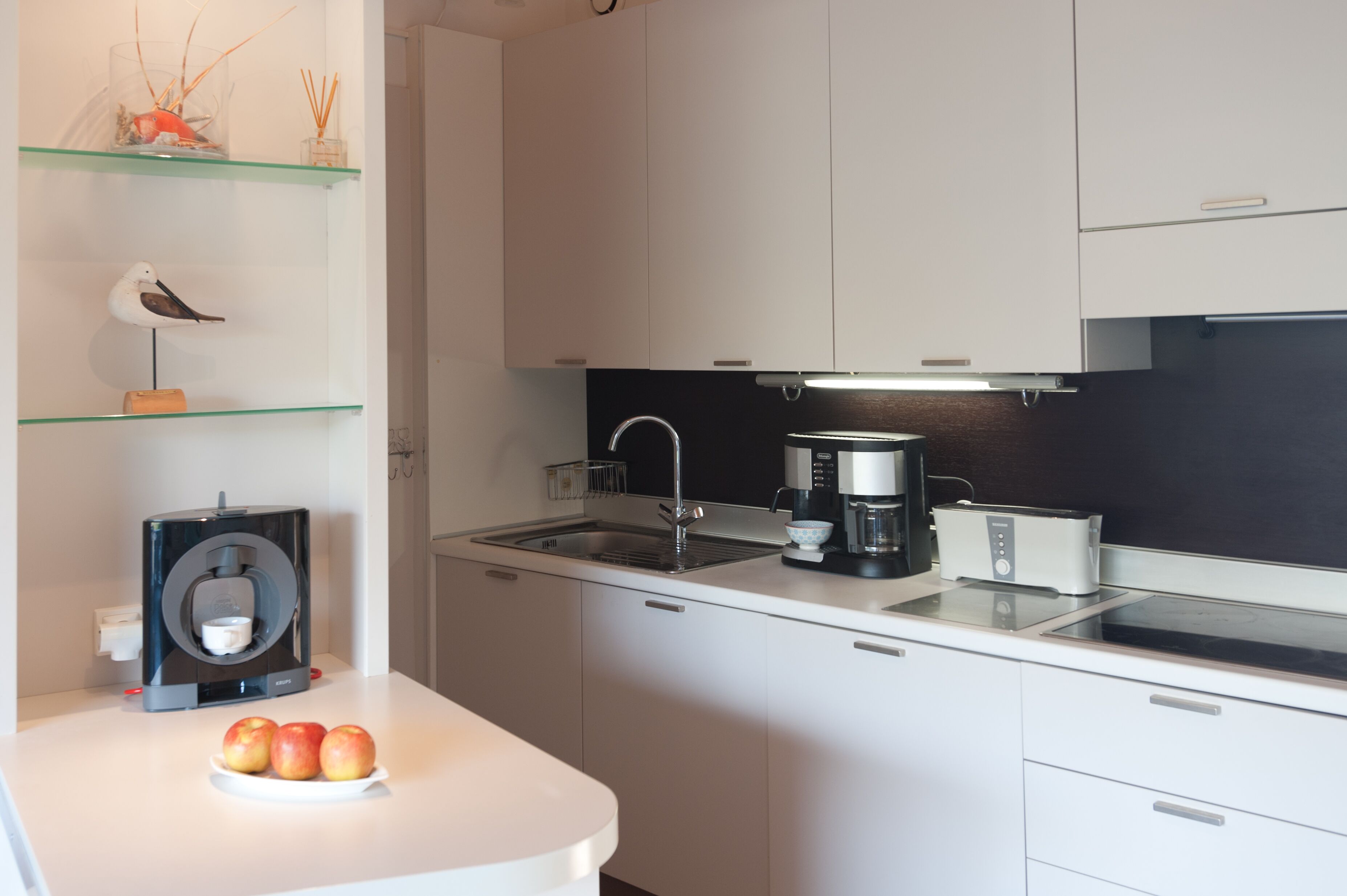 Una cocina moderna con armarios blancos, encimera negra, una cafetera y un plato de manzanas.