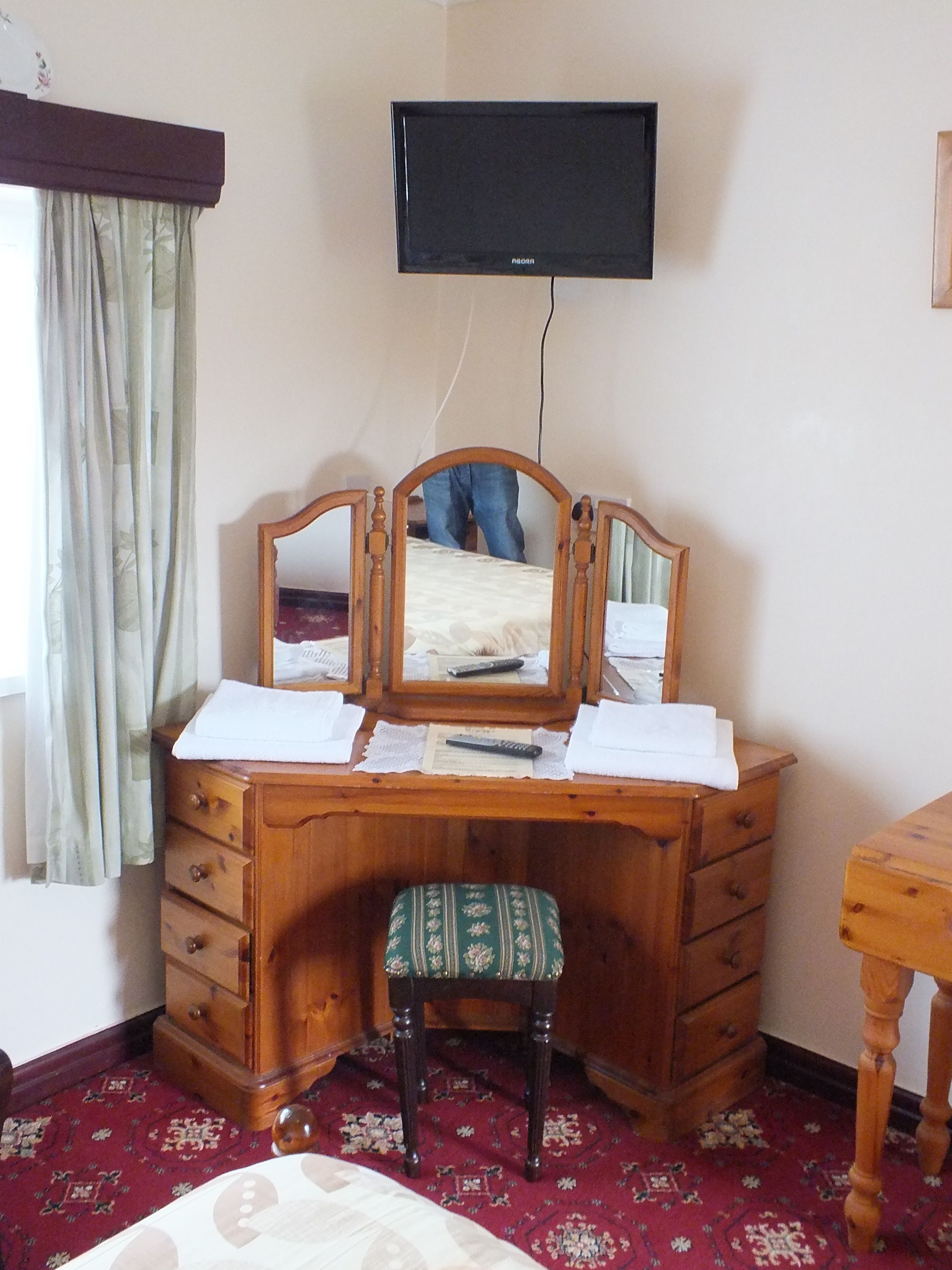 A wooden desk with a mirror, a television mounted above it, and a small stool.