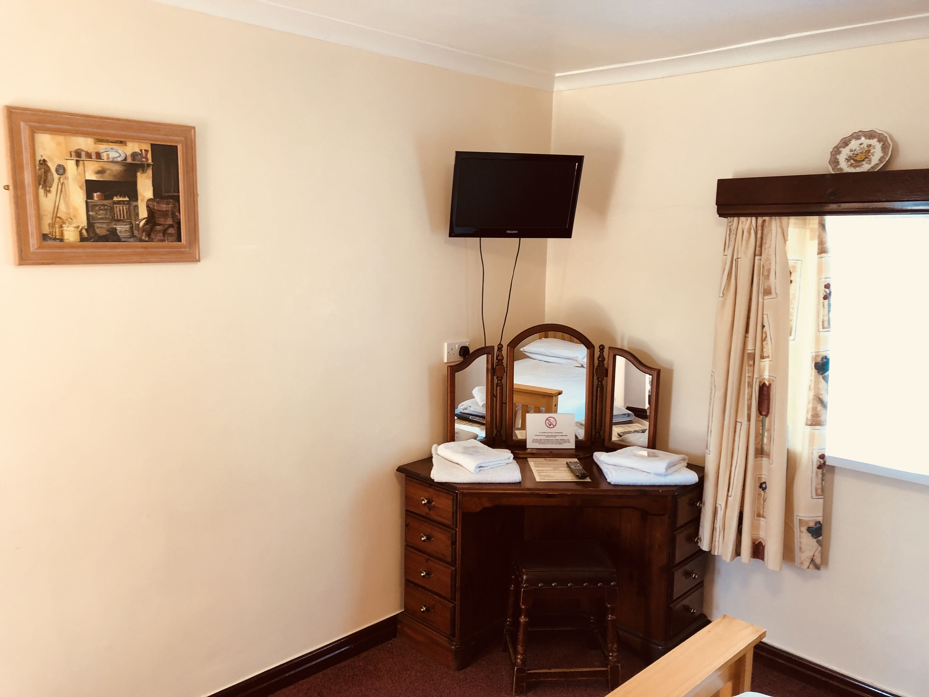 A room with a wooden vanity, a flat-screen TV mounted on the wall, a small stool, and a window with curtains.
