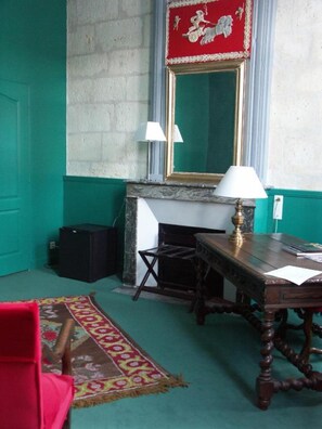 Una habitación con alfombra verde, un espejo con marco rojo y dorado, una chimenea, una mesa de madera con una lámpara y una silla roja.