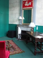 A room with a green carpet, a red and gold framed mirror, a fireplace, a wooden table with a lamp, and a red chair.