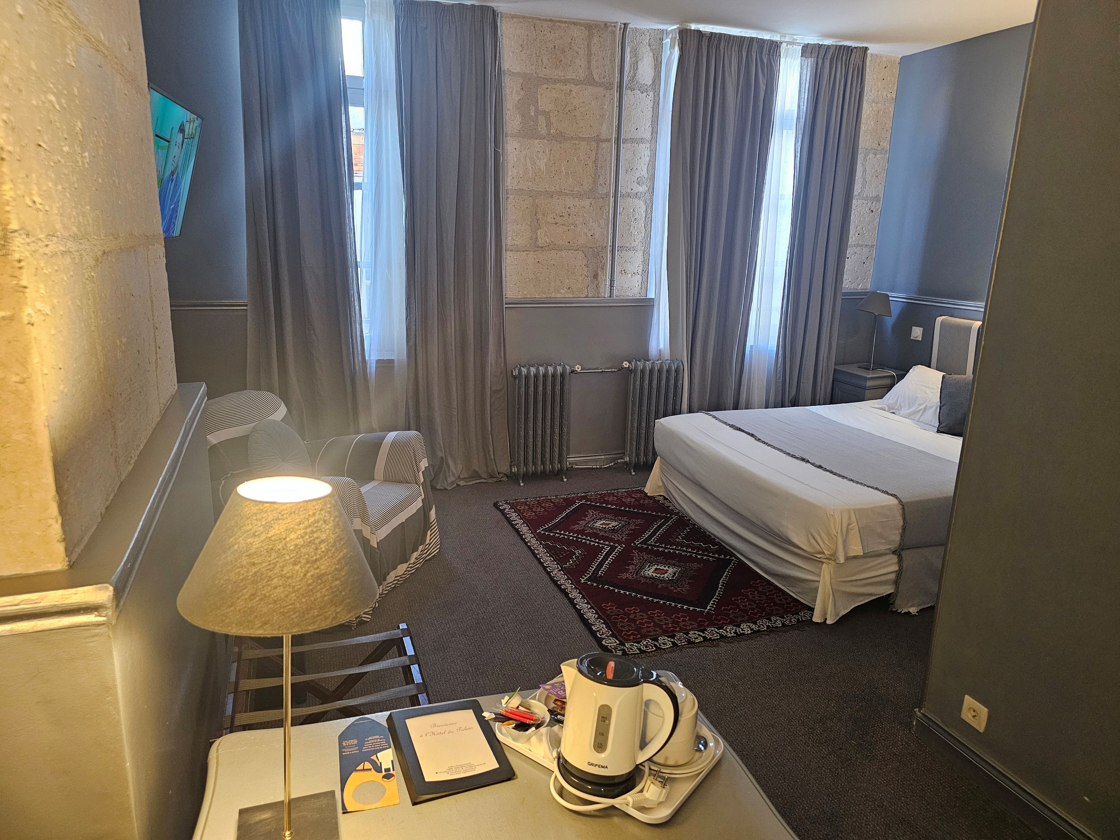 Habitación de hotel con cama, escritorio con lámpara, radiador y ventana con cortinas.