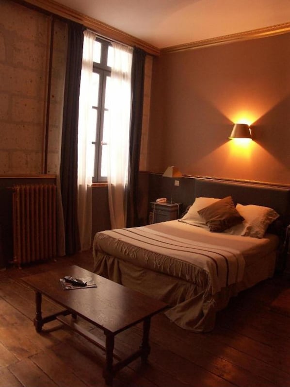 Una habitación con cama, mesita de noche, mesa y ventana con cortinas.