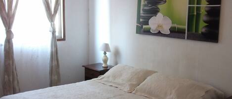A bedroom with a bed, a nightstand, a lamp, a window with curtains, and a wall painting.