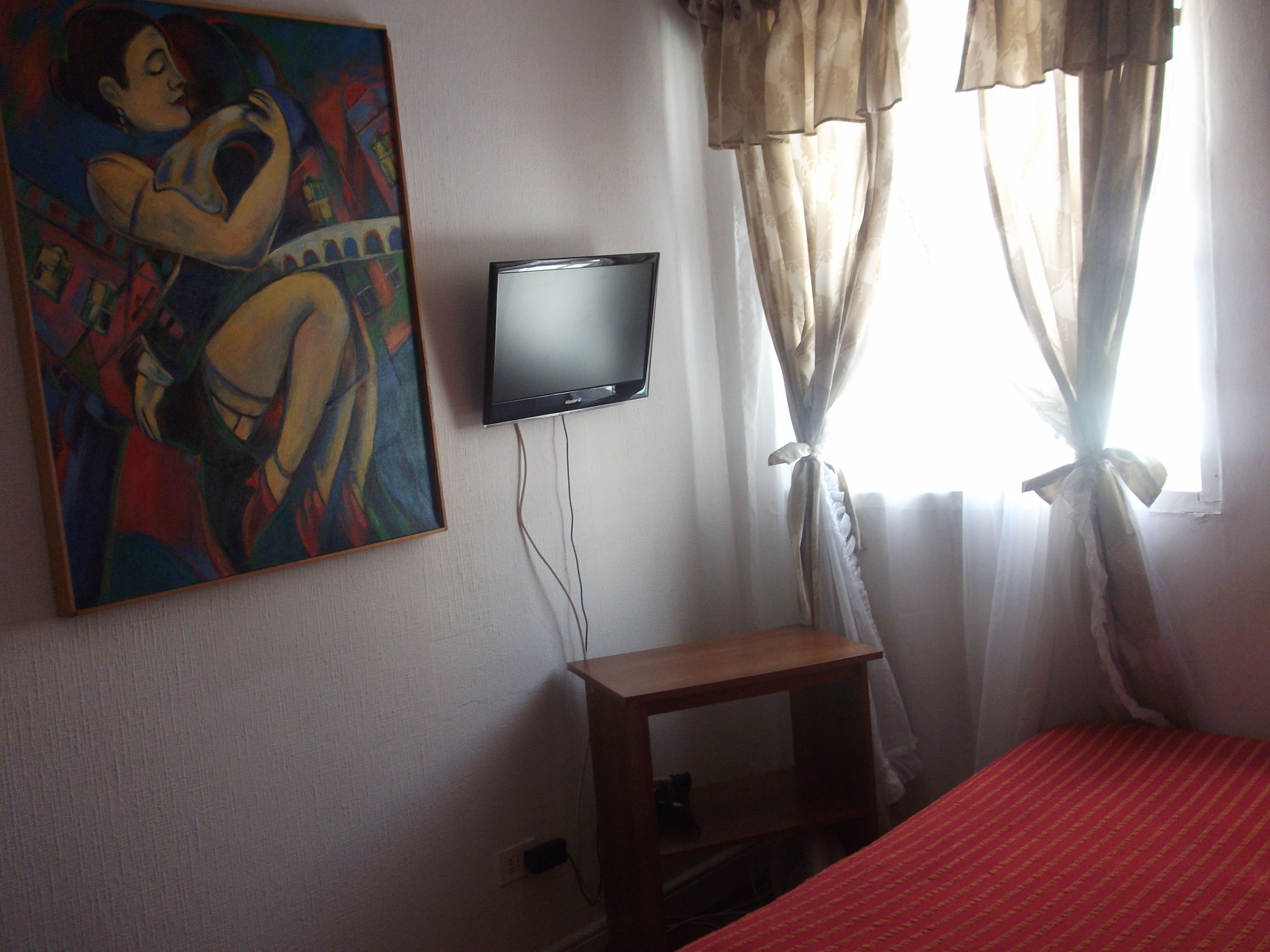 A room with a bed, a television mounted on the wall, a painting, and a window with curtains.