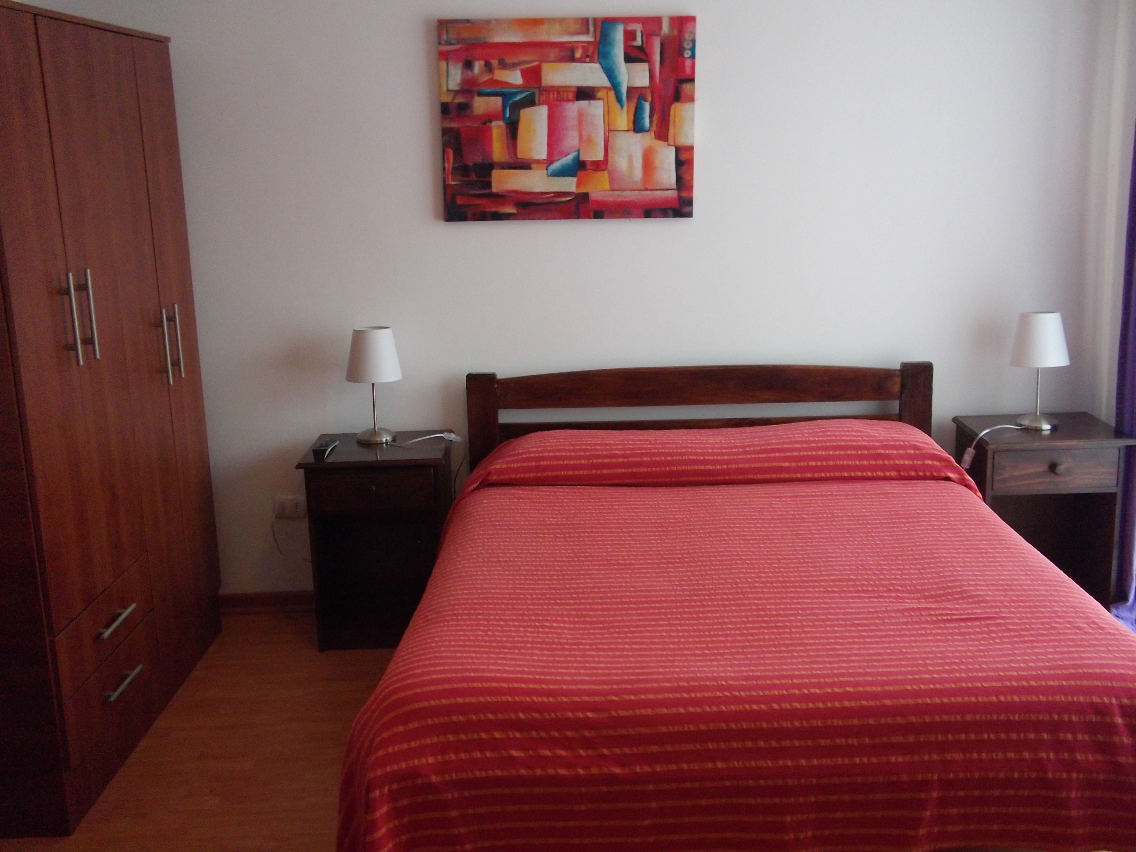 A bedroom with a bed, a wooden wardrobe, two bedside tables with lamps, and a colorful abstract painting on the wall.